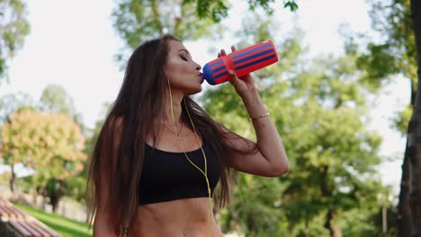 Young-fitness-woman-drinking-water-from-plastic-bottle-after-morning-jogging,-listening-to-the-music-and-drinking-water-from-the