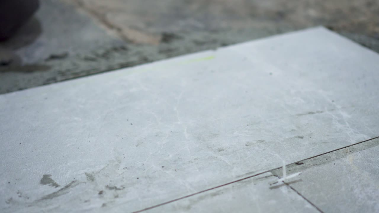 A close look at the construction tiler worker laying down the tile on cemented floor. Professional worker laying tiles on floor at construction site close-up slow motion.