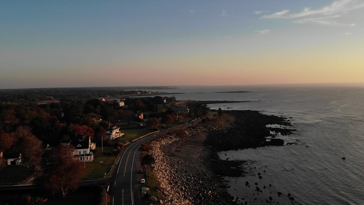Drone footage captures a scenic coastal road winding along rocky shoreline with elegant white New England style homes during golden hour sunset.