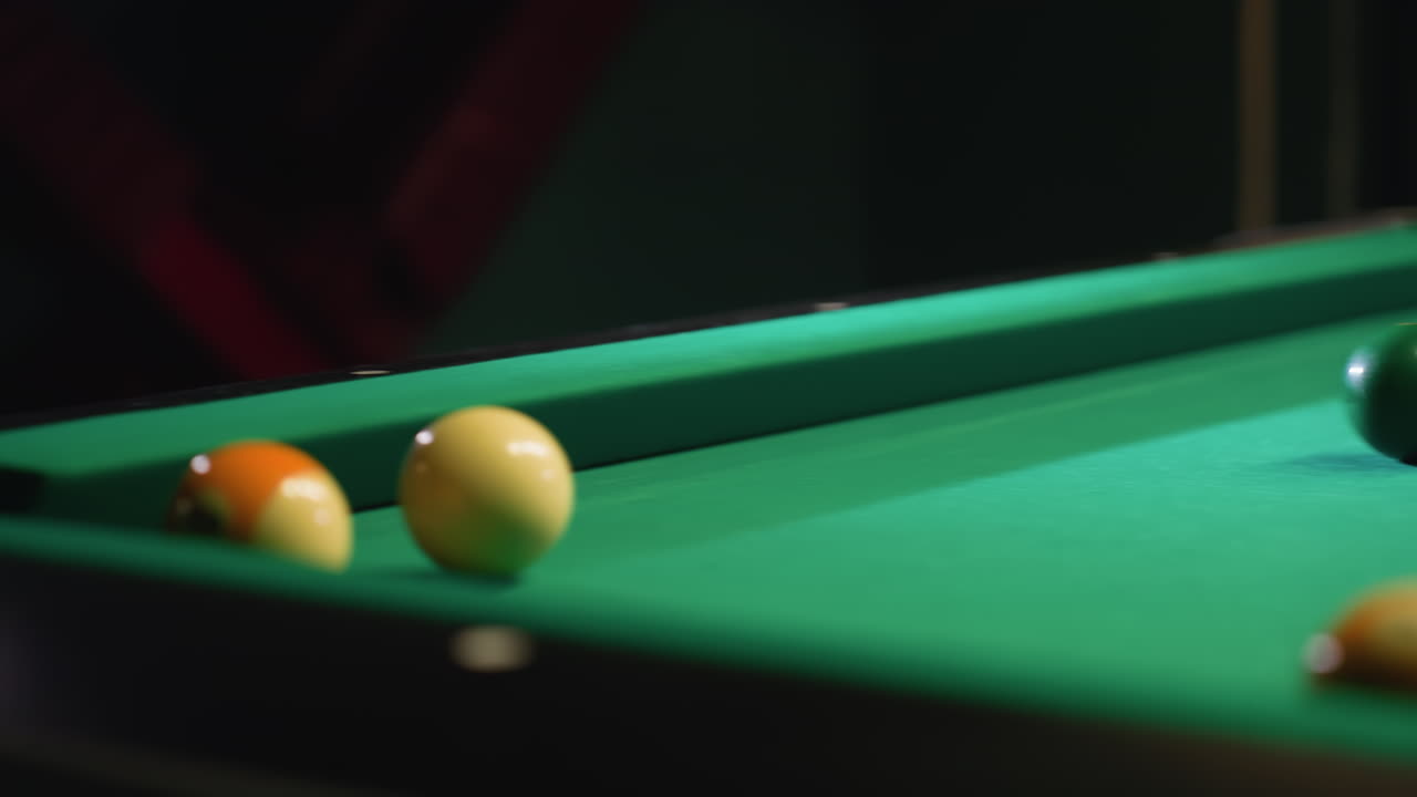 Cue stick strikes white billiard ball, sending it toward orange ball on green pool table. Blurred billiard balls in background enhance depth, highlighting movement, precision