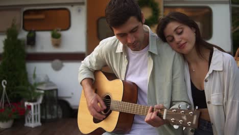 romántica, hermosa pareja sentada al aire libre delante de su furgoneta y el hombre tocando la guitarra para la novia. al aire libre. chica escuchando música, apoyándose en su hombro