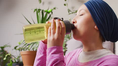 video of tranquil biracial woman in hijab drinking from water bottle at home