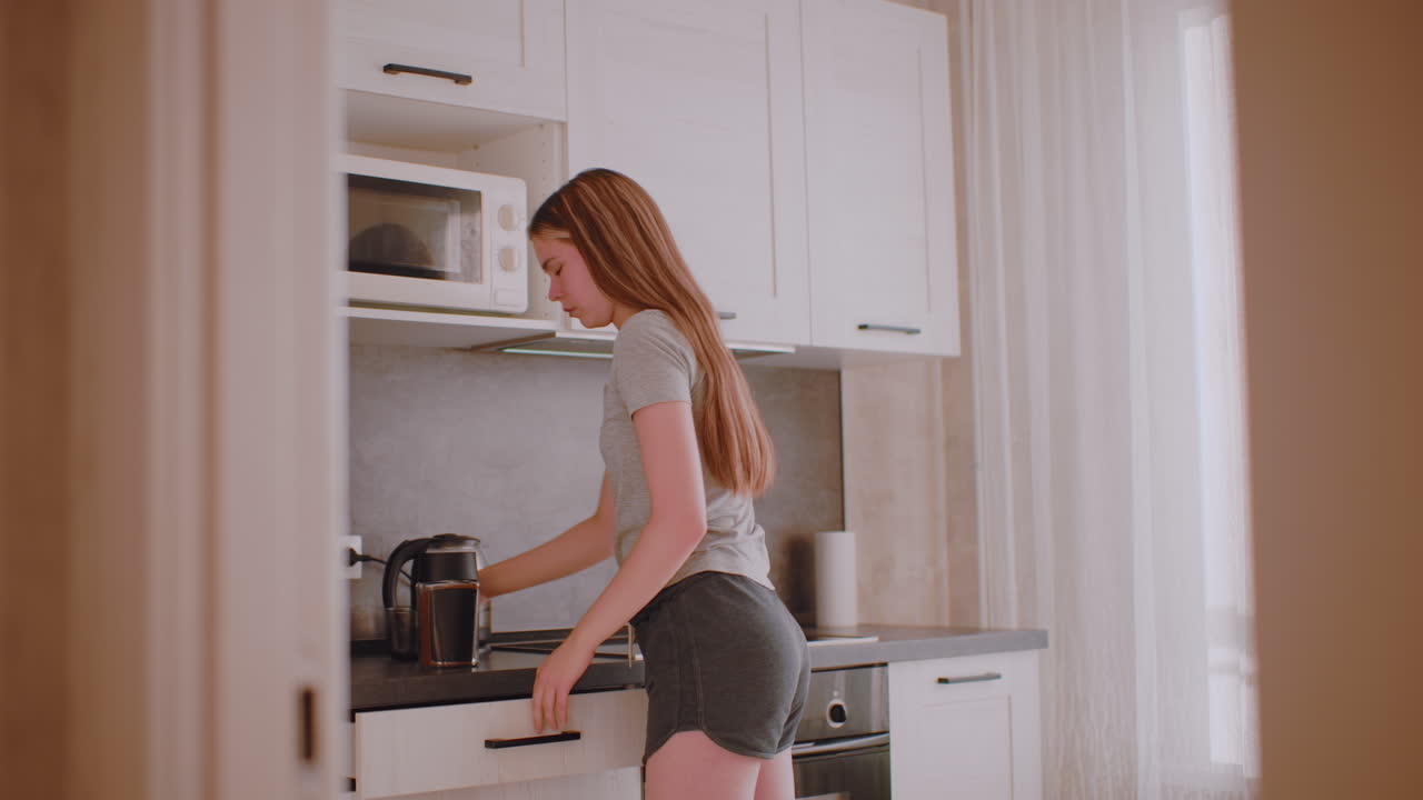 Freelancer prepares for work in kitchen, taking beverage container from cabinet, placing it on counter, bringing cup forward, opening drawer for spoon under natural daylight atmosphere