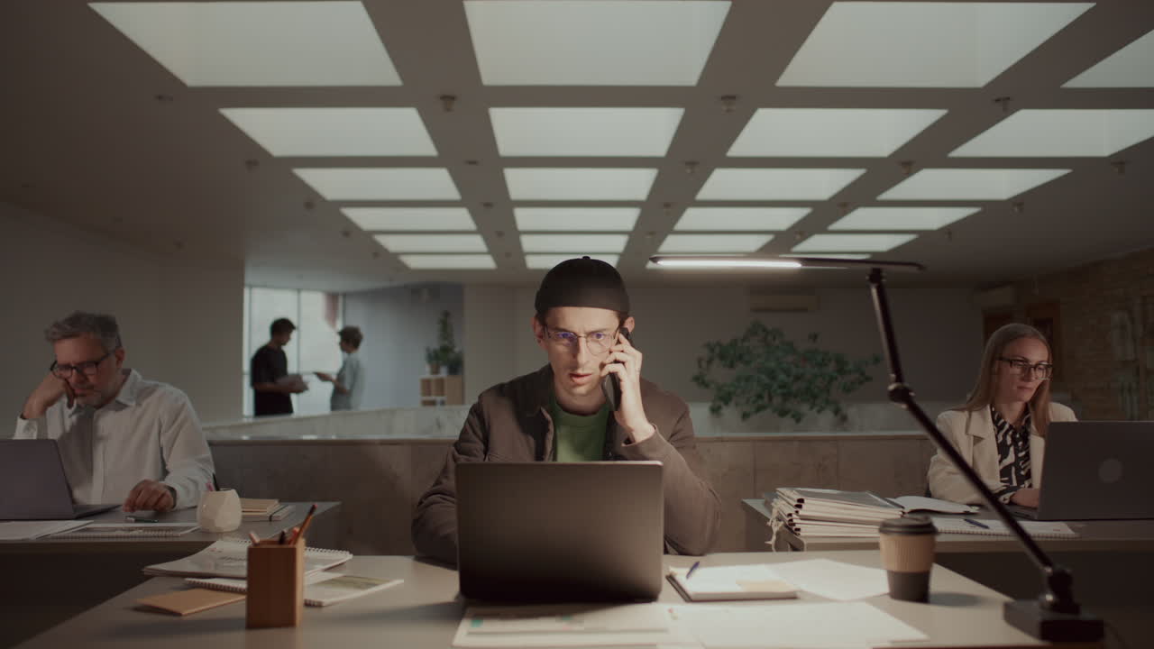 Man Using Laptop and Speaking on Phone at Desk in Open-Plan Office