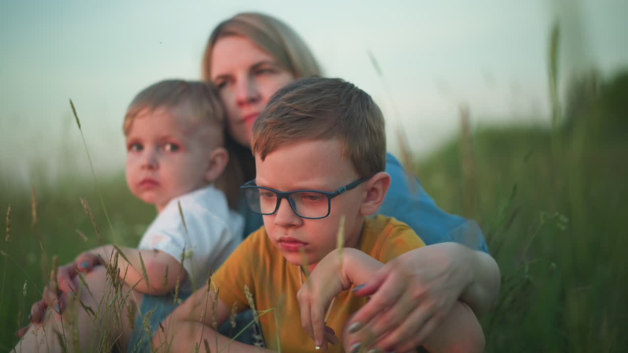 A close-up front view of a mother in a blue gown sitting in a field with her two children, one in a yellow top and the other in a white top. They are all gazing at their surroundings