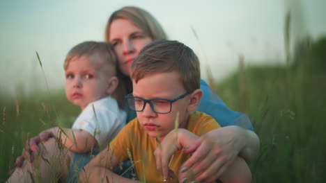 una vista frontal de cerca de una madre con una túnica azul sentada en un campo con sus dos hijos, uno con una camiseta amarilla y el otro con una blanca. todos están mirando a su alrededor