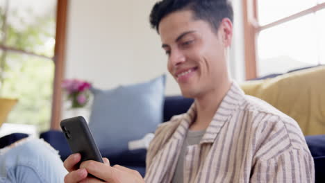 Hombre-Birracial-Feliz-Sentado-En-El-Suelo-Usando-Un-Teléfono-Inteligente-En-La-Sala-De-Estar,-Cámara-Lenta