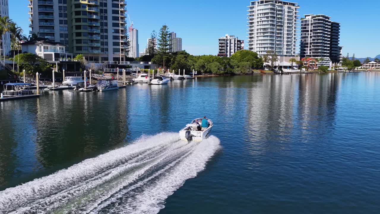 Aerial footage of a speedboat gliding along a calm waterway, leaving a wake, surrounded by modern high-rise buildings under clear daylight