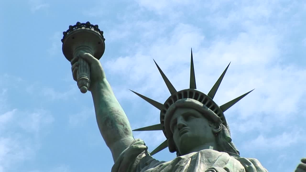 La Statua della Libertà con la torcia levata in alto si erge contro un cielo blu pieno di nuvole sottili.