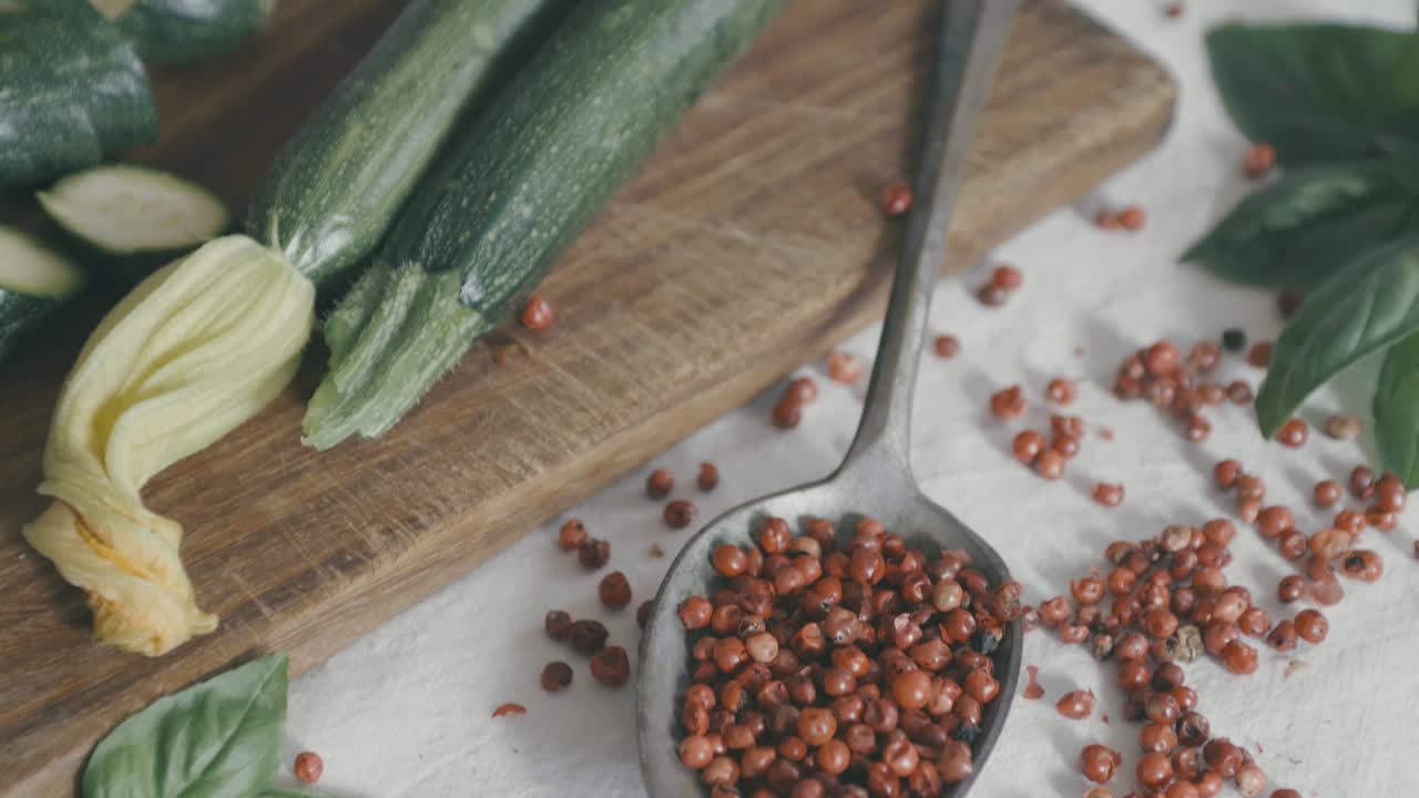 incline de abajo hacia arriba sobre una mesa en la que hay hojas de albahaca, pimiento rojo y calabacines, una mano llega y toma un calabacín