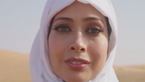 close up portrait of a beautiful muslim woman in white hijab standing in a windy desert and smiling at camera