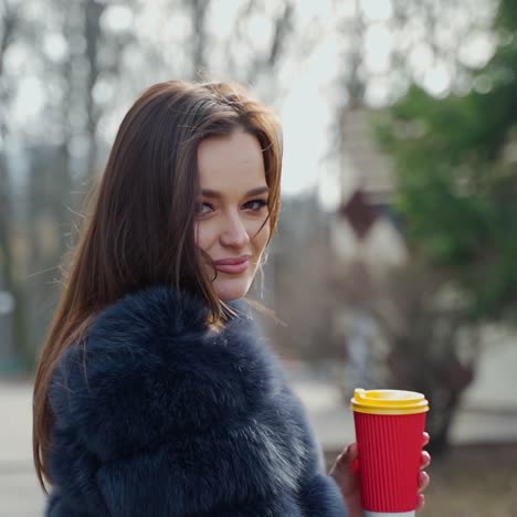 attractive woman holds coffee in a plastic cup. portrait of a beautiful girl in fur coat calling someone with her hand in the city park. slow motion.
