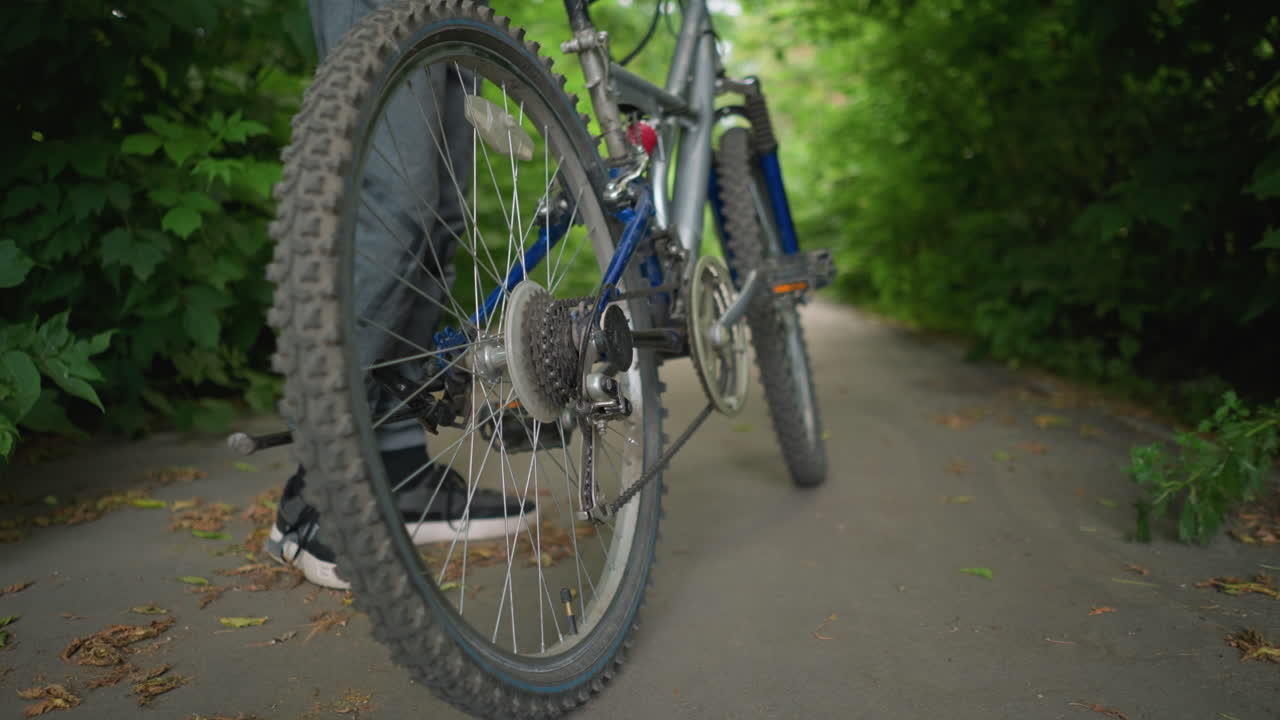 primer plano de alguien subiendo a la bicicleta, colocando su pie derecho en el pedal en un camino pavimentado rodeado de exuberante vegetación, hojas secas están esparcidas por el suelo