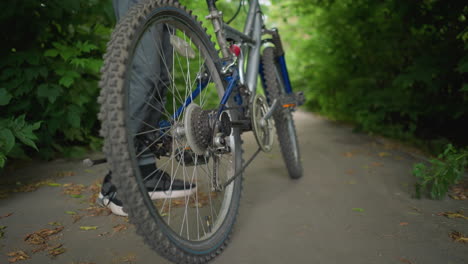 close-up of someone climbing bicycle, placing their right foot on the pedal on a paved path surrounded by lush greenery, dry leaves are scattered across the ground