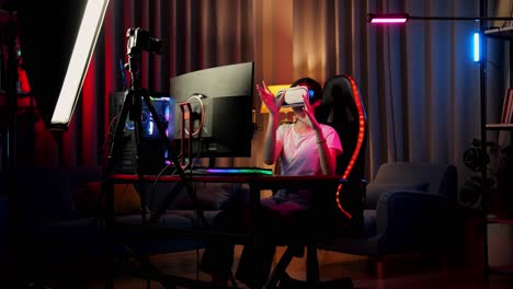 asian girl streamer playing game experience virtual reality in futuristic goggles. player touching air panel. personal computer on desk illuminated by rgb led strip light