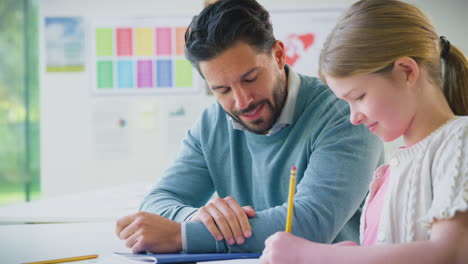 Profesor-Con-Estudiante-En-El-Aula-De-La-Escuela-Sentado-En-El-Escritorio-Escribiendo-En-Un-Libro-Juntos