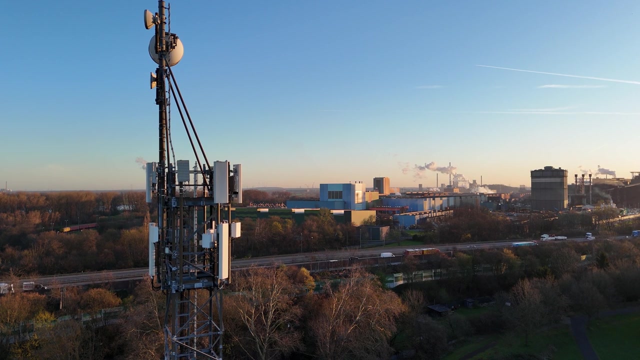 Locked drone view shows a telecom tower beside the motorway, with passing vehicles and industrial background from a frontal angle.