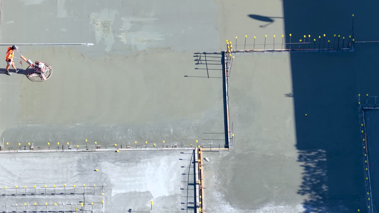 Worker uses power trowel to smooth fresh concrete slab, aerial view, strong sunlight, sharp shadows