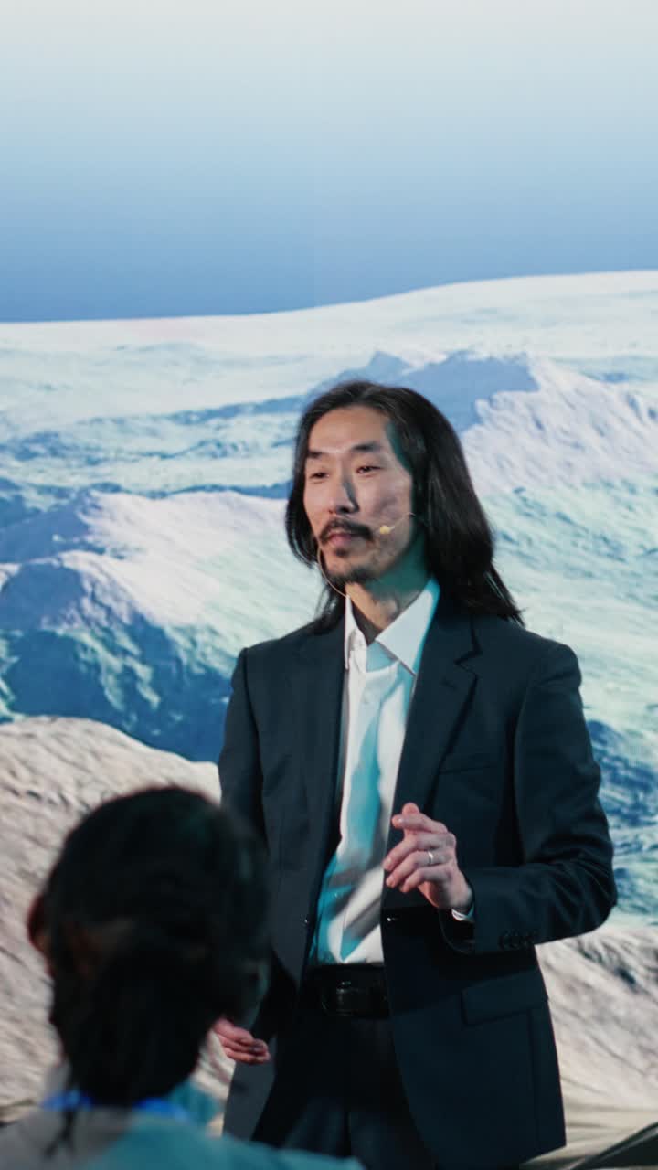 Video verticale: oratore asiatico tiene una presentazione sull'energia eolica su un palco con turbina eolica