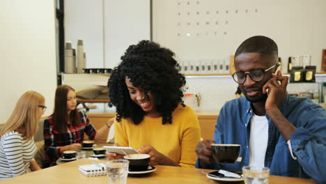 Hombre-Afroamericano-Hablando-Por-Teléfono-Y-Mujer-Afroamericana-Viendo-Un-Video-En-Una-Tableta-Sentada-En-Una-Mesa-En-Un-Café