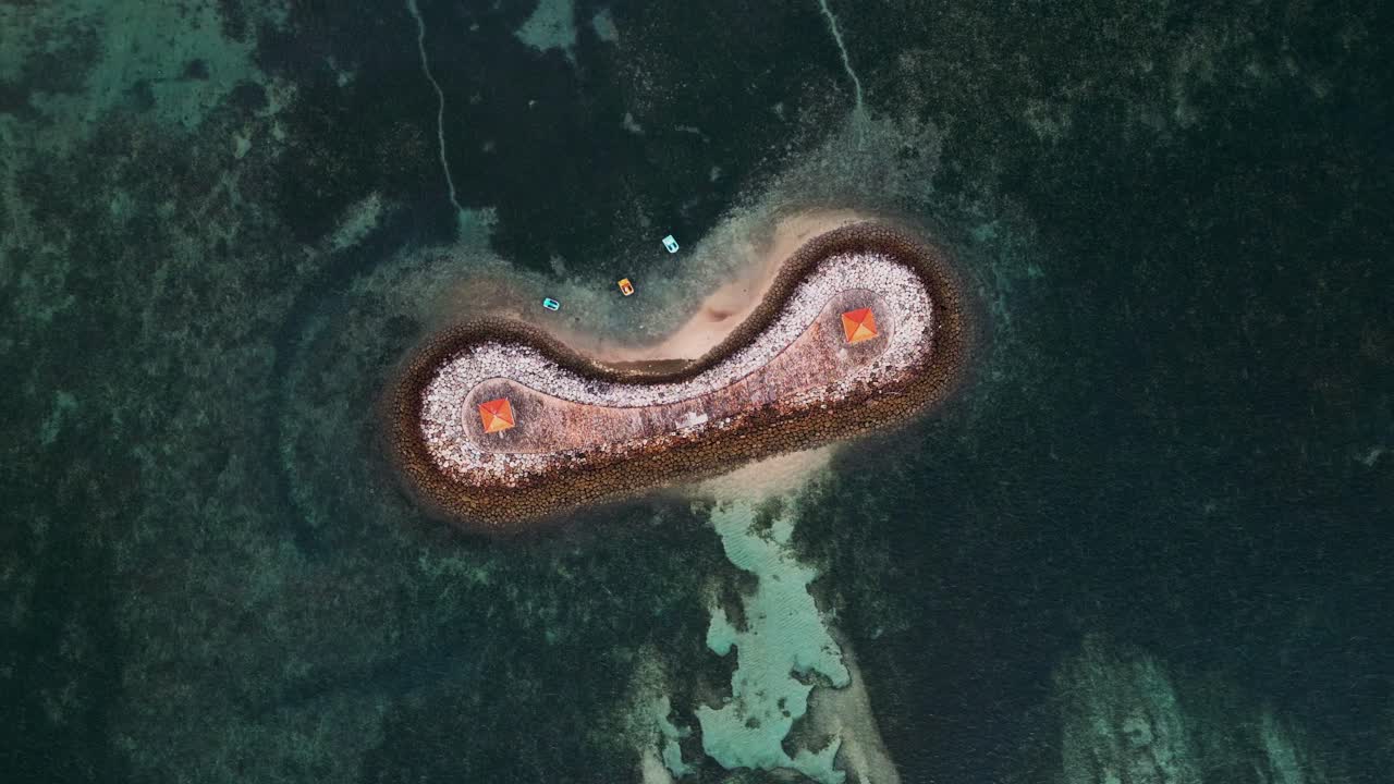 Aerial drone footage of Sanur Bali showing a curved stone wave breaker with a red roof pavilion, turquoise waters, fishing boats, and calm coastal scenery along the island shoreline