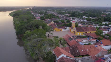 the drone glides over the magdalena river, rolling and yawing right to unveil the majestic front of santa barbara church. the golden light casts soft shadows on the church’s intricate facade. mompox