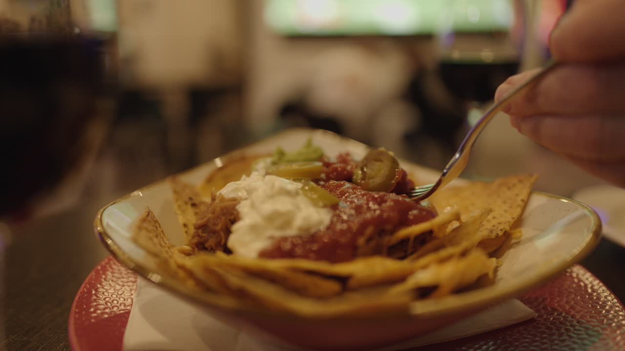 Hand scoops nachos with salsa, cream, and jalapenos in dimly lit bar with television