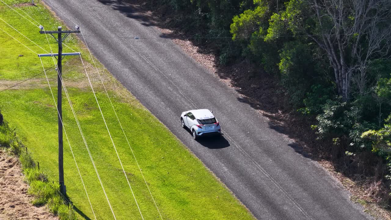 Drone captures a car driving through a lush, green rural landscape under bright sunlight