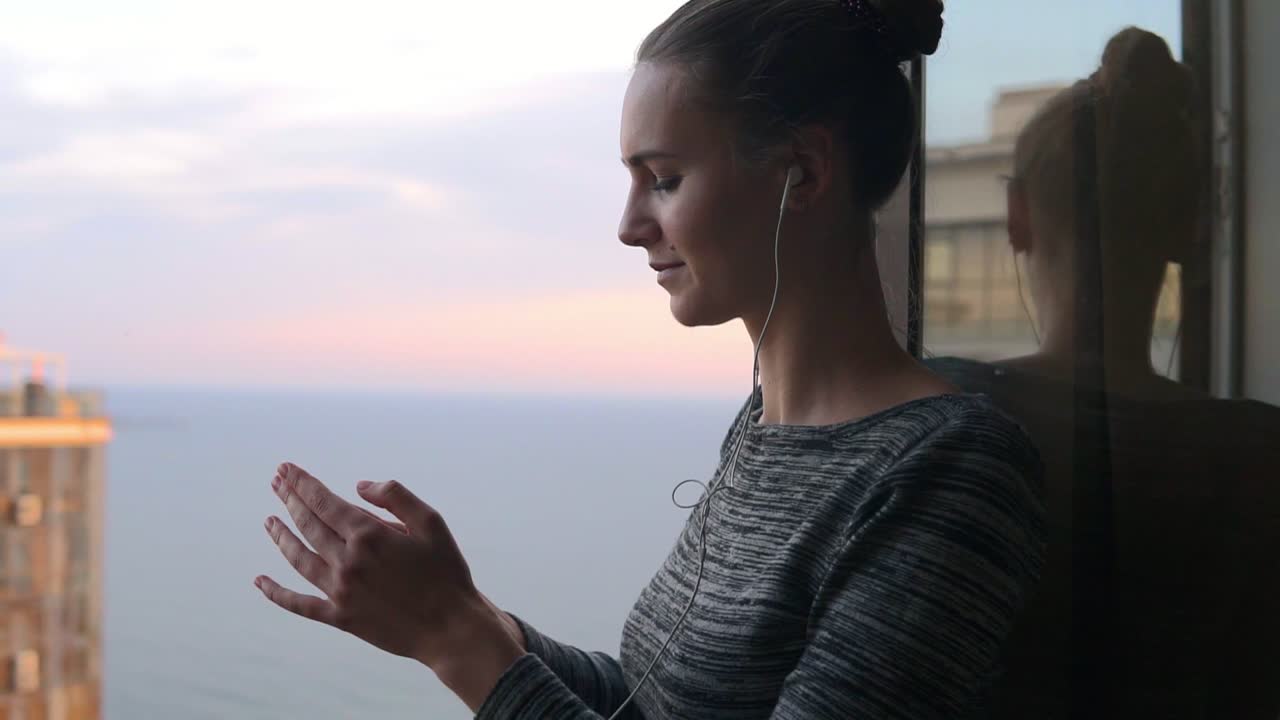 niña sonriente de pie junto a la ventana durante un día soleado y escuchando música con auriculares usando teléfono móvil