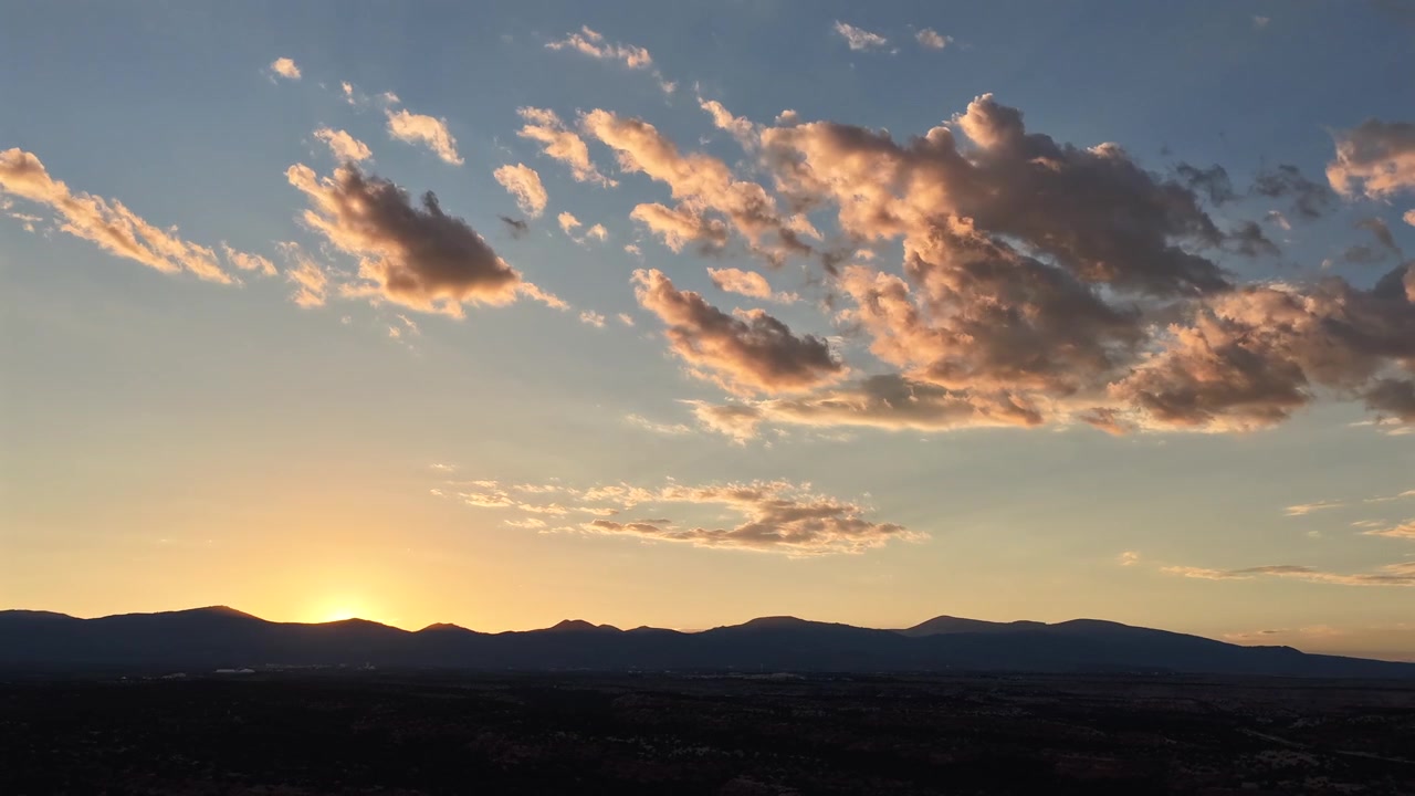 4K drone timelapse capturing the sun disappearing behind a dark, silhouetted southwest mountain range. Warm, beautiful clouds dance in place against a serene and fading blue evening sky.