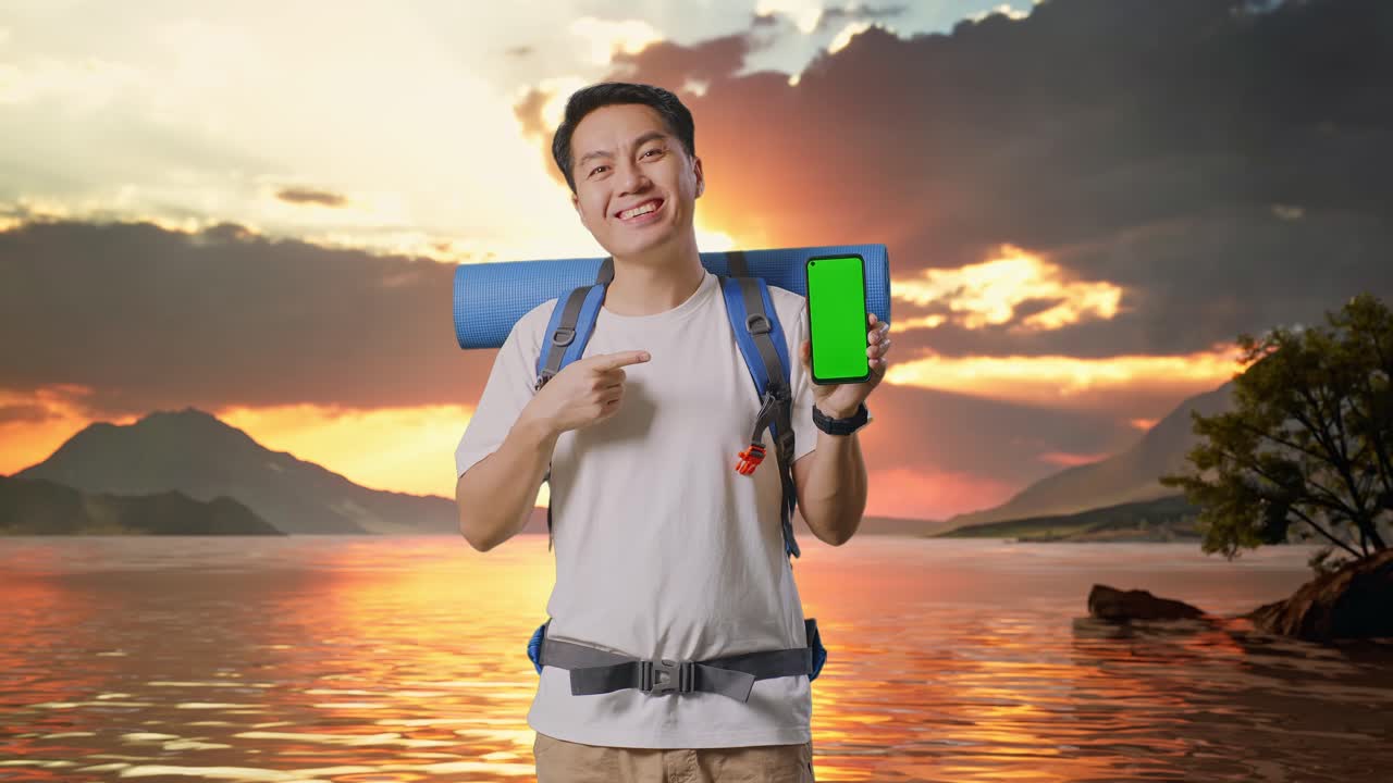 Asian Male Hiker With Mountaineering Backpack Smiling And Pointing To Mock Up Green Screen Smartphone At A Lake