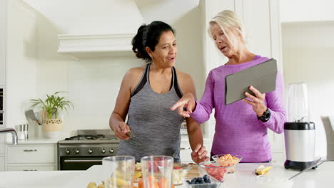 two happy diverse senior women preparing fruits using tablet in kitchen, slow motion