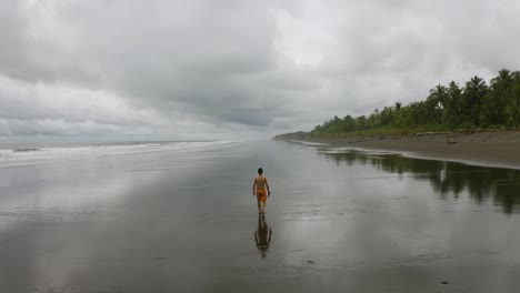 Antena-Sigue-A-Un-Joven-Desde-Atrás,-Caminando-En-Una-Playa-Vacía-En-Nuqui,-Colombia