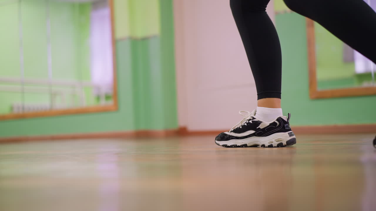 Side leg view of girl in black leggings and canvas sneakers bouncing on polished wooden floor during workout session, capturing movement, rhythm, and energy with blurred background of indoor studio