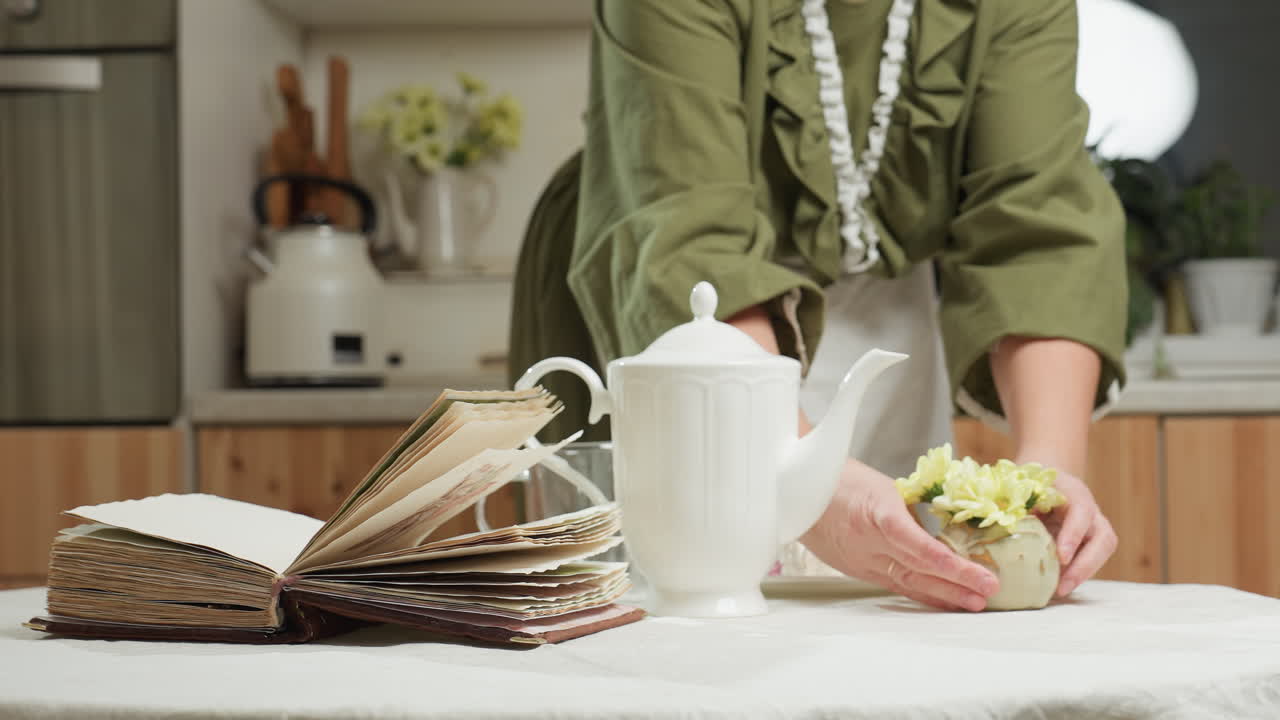 Woman prepares breakfast table by placing flower vase next to white ceramic teapot and open book while wearing green outfit in cozy kitchen, then sits down and gently lifts glass cup