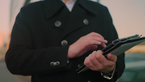 close-up of person in black coat operating tablet near glass building, reflecting parked cars and blurred figures in glass, showcasing modern city environment