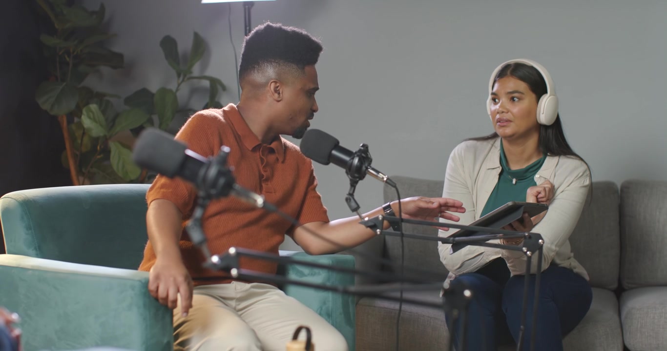 Conduttori afroamericani: un uomo si china, una donna indica e tocca un tablet vicino ai microfoni mentre registrano un podcast.
