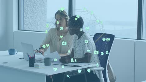 Animation-of-connected-icons-forming-globe,-diverse-female-coworkers-discussing-reports-on-laptop