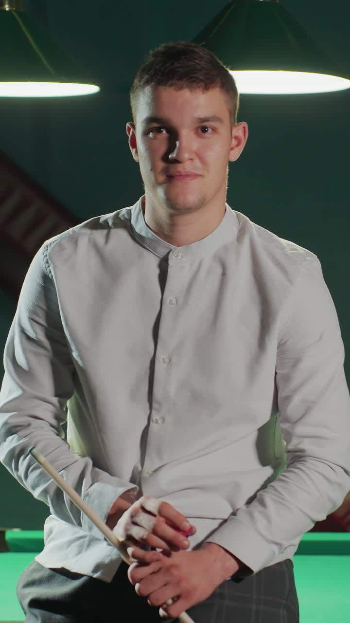 Giovane uomo in camicia bianca e pantaloni grigi seduto su un tavolo da biliardo verde, che tiene in mano una stecca da biliardo e sorride. Le palle da biliardo e l'attrezzatura da biliardo sono visibili sullo sfondo sotto una calda illuminazione.