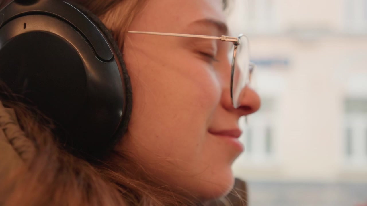 Donna bianca che ascolta musica con le cuffie, godendosi la vista dalla finestra, primo piano di profilo con occhiali e cuffie over-ear, atmosfera cinematografica rilassata che trasmette un suono ambientale, bokeh morbido e un'atmosfera di pendolarismo urbano tranquillo.