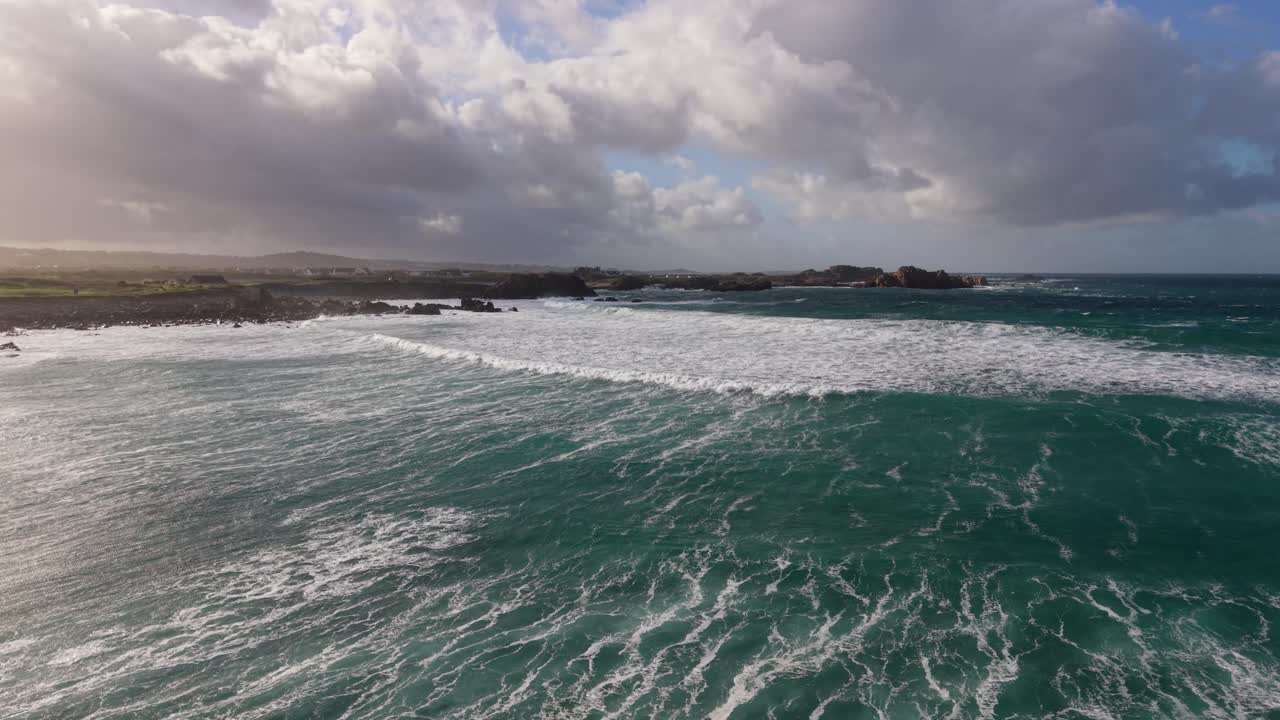 Vuelo bajo al anochecer sobre una extensa playa que se adentra en el mar con olas blancas espumosas rompiendo en la orilla, con luz mágica y cielo amenazante al atardecer, con una bandada de gaviotas volando.