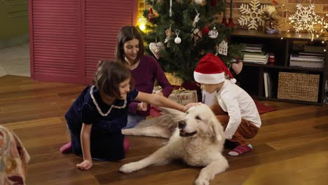 Tres-Niños-Acariciando-A-Un-Perro-Golden-Retriever-Y-Riéndose-En-El-Suelo-Bajo-Un-árbol-Decorado-De-Año-Nuevo,-En-Cámara-Lenta