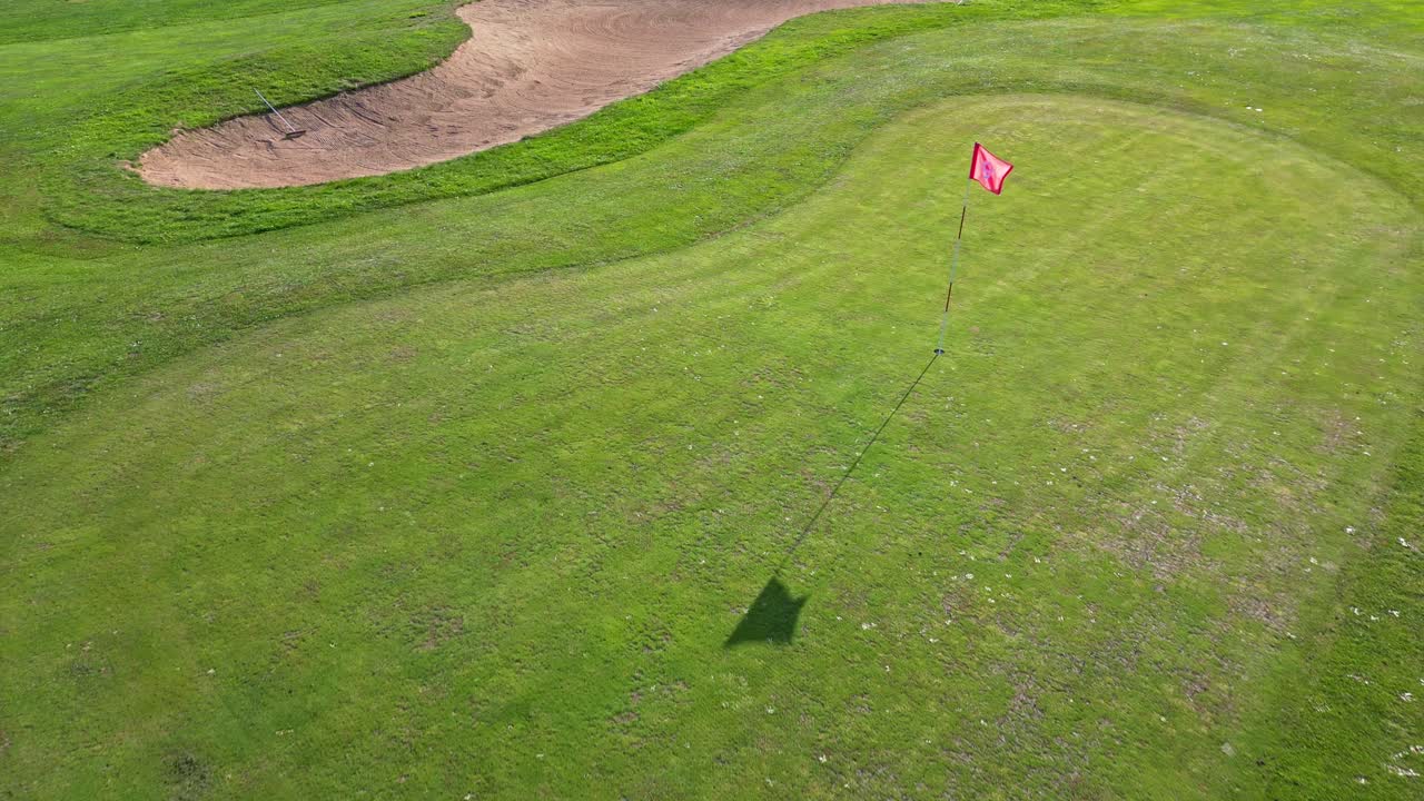 Drone moving forward in a near top-down view over a golf course, showing the flag, its shadow on the grass and a sand bunker
