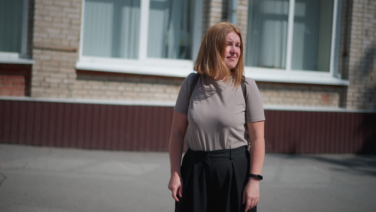 Melancholic girl stands under bright summer sun near building, looking around for someone and checking time with fitness bracelet, appearing exhausted and thoughtful, her long shadow visible