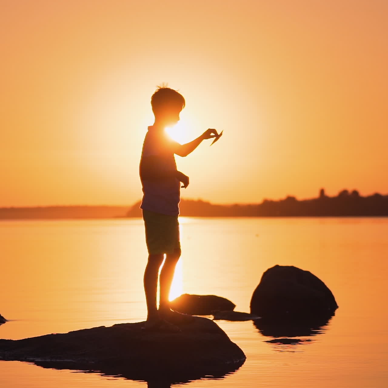 Silhouette of a boy with a paper plane at sunset. Child is standing on a stone on the orange river background. Bright rays of evening sun shines on a boy.