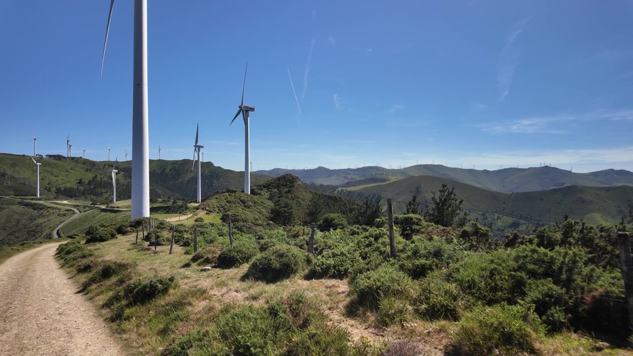 Turbinas eólicas erguendo-se de encostas verdes de montanhas sob um céu azul claro.