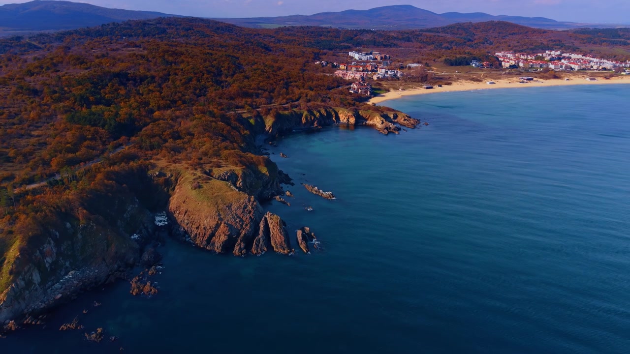 Veduta aerea di una costa rocciosa con colline boschive autunnali e spiaggia del resort al crepuscolo. La calda luce della sera rivela una costa rocciosa autunnale dove una caletta appartata si apre di fronte a edifici del resort in lontananza