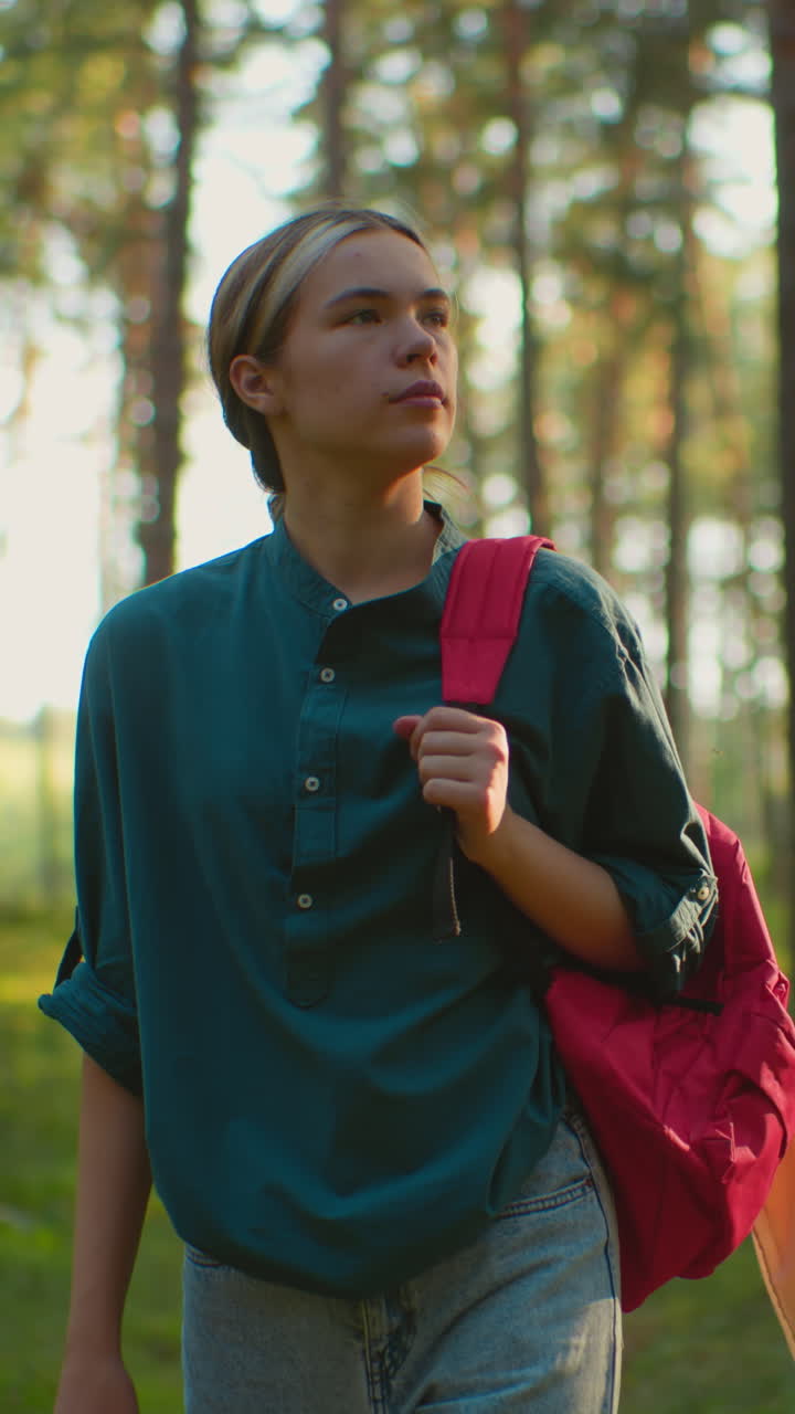 Woman walking through tranquil forest, carrying red backpack and gazing into distance with serene expression, surrounded by tall trees, sunlight casting warm glow behind her