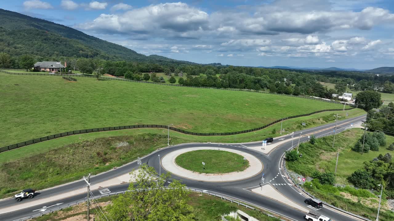 Aerial view of modern busy roundabout in rural Virginia, surrounded by green fields, treesand distant mountains under partly cloudy sky. Scenic countryside traffic and infrastructure landscape in USA