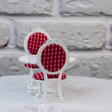 carved wooden toy table and two chairs at both sides rotating. pretty furniture for a dolly house. white table and red-and-white chairs against brick wall backdrop